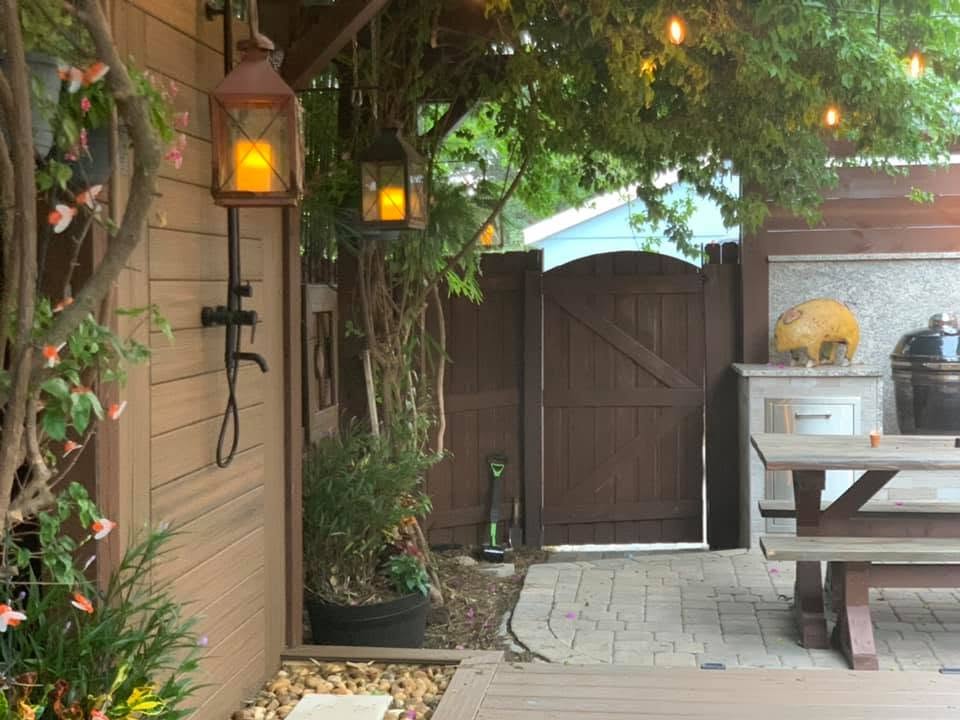 A backyard patio features an outdoor shower, hanging lanterns, and a wooden gate.