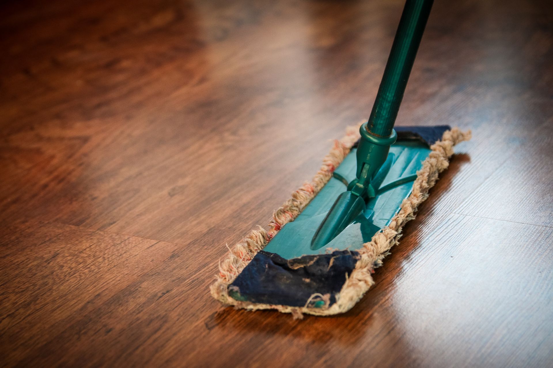 A green flat mop cleaning a dark wood-look vinyl floor.